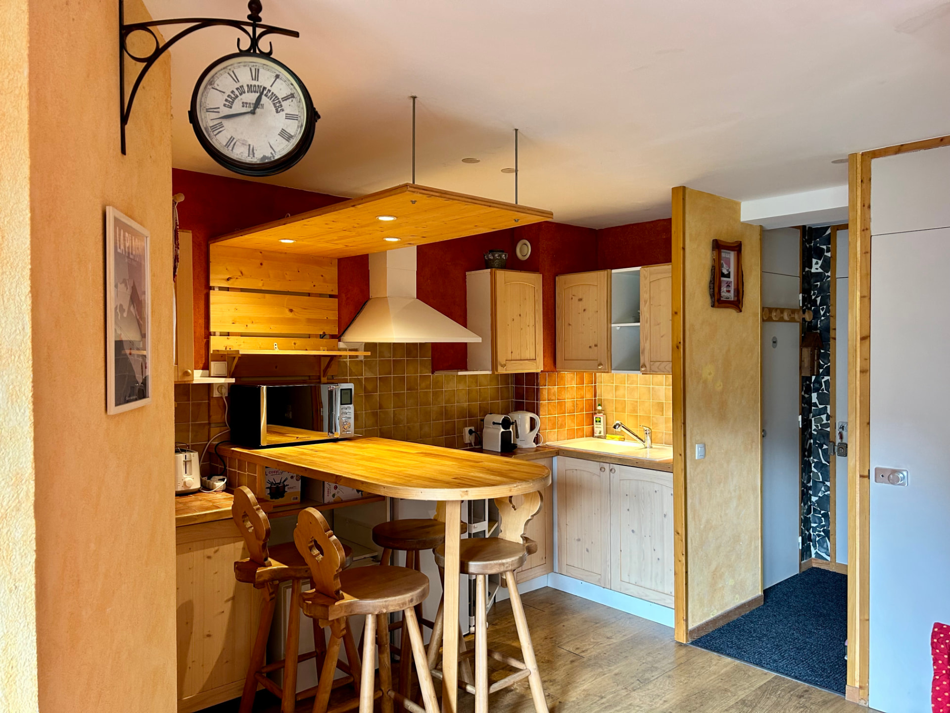 Coin cuisine et bar en bois avec tabourets, horloge de gare et entrée de l'appartement